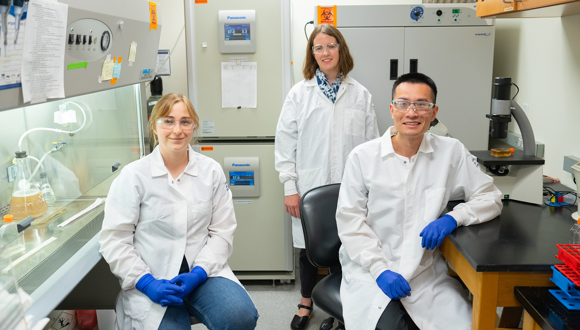Three researchers wearing white lab coats, blue gloves, and protective glasses pose in a biology lab. Two are seated—one woman on the left and one man on the right—and a third woman stands behind them. Lab equipment and supplies are visible in the background, including incubators, a biosafety cabinet, and a microscope.