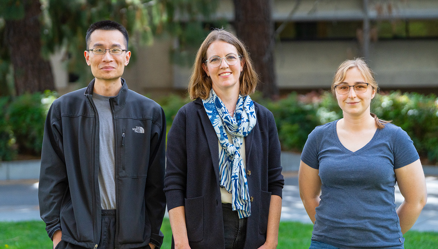 Three researchers—two graduate students and a faculty member—stand outdoors, smiling at the camera with trees and a building in the background.