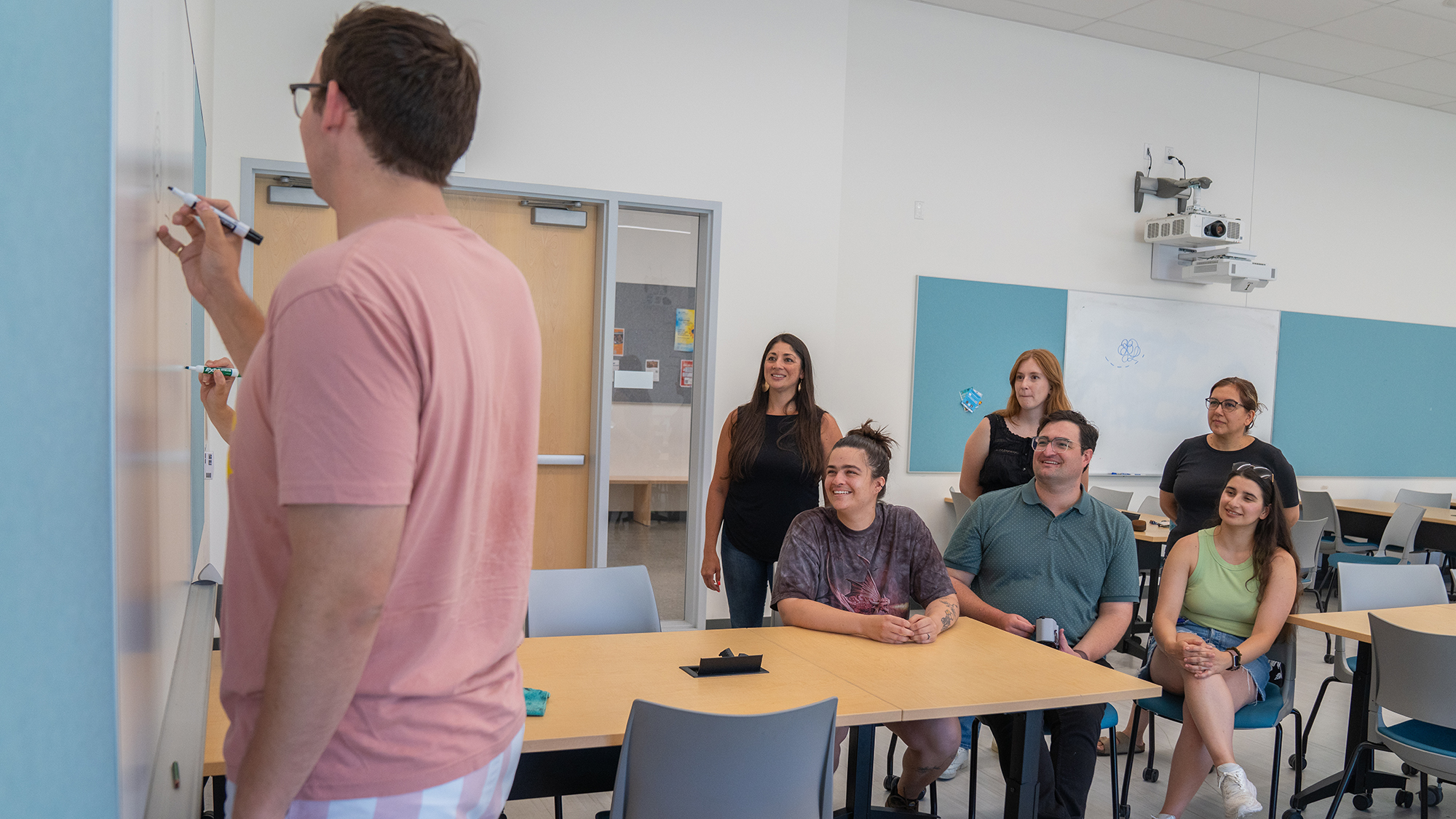 Graduate students sit at tables and watch a classmate write on the whiteboard during a group activity in a bright classroom.