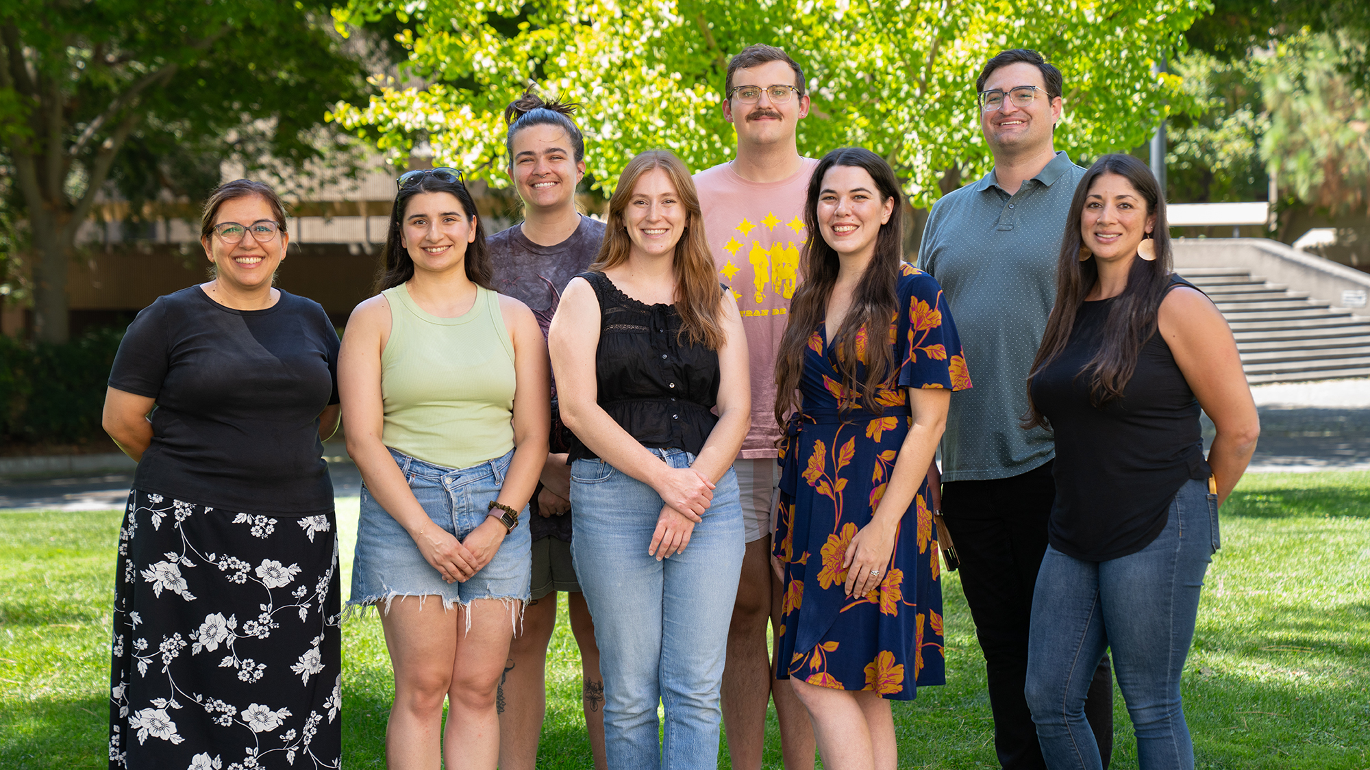 Eight people stand together outdoors in a group portrait, smiling in front of green trees on a sunny day.