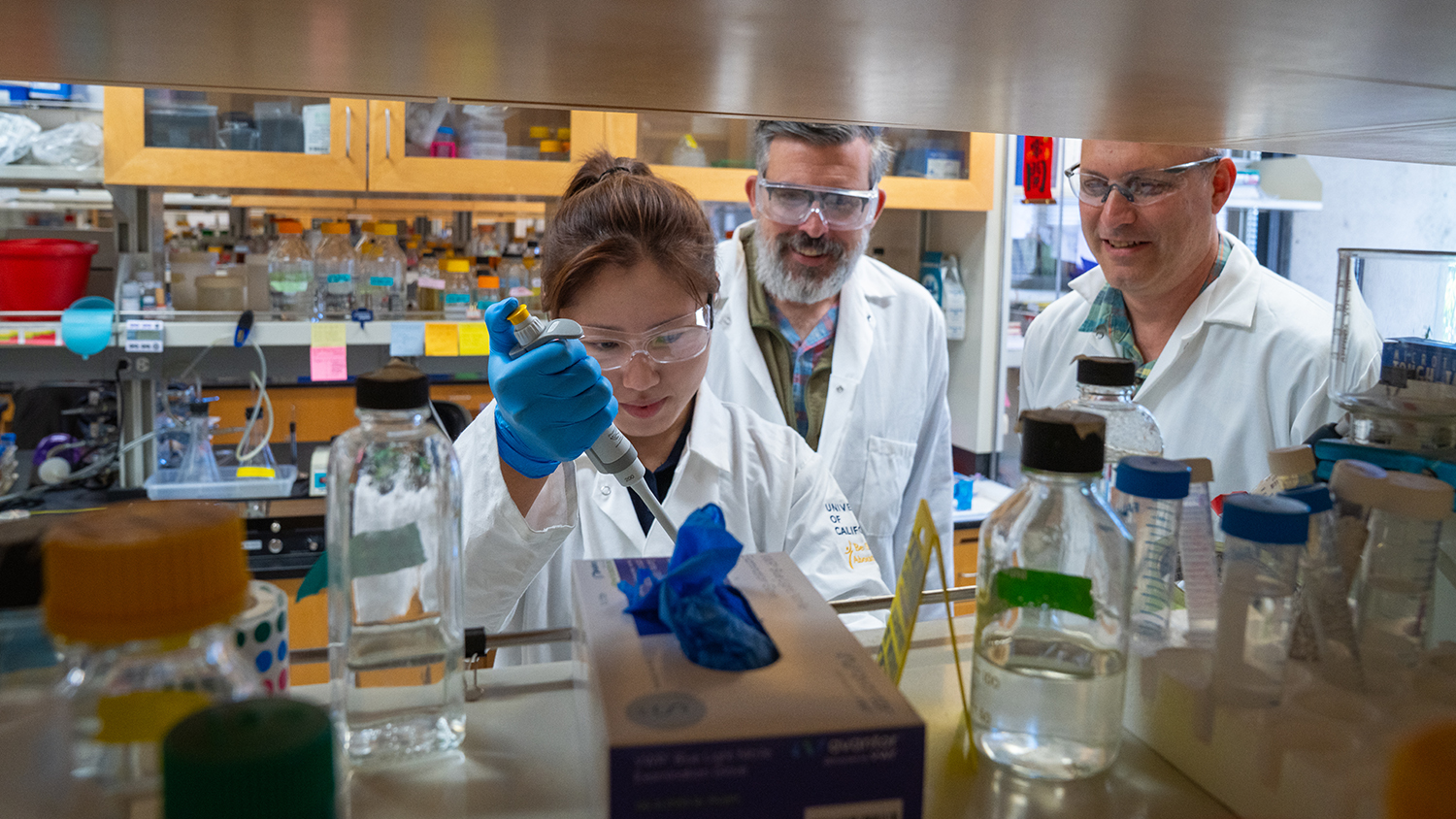 Graduate student pipettes a sample in the lab while two professors observe from behind.
