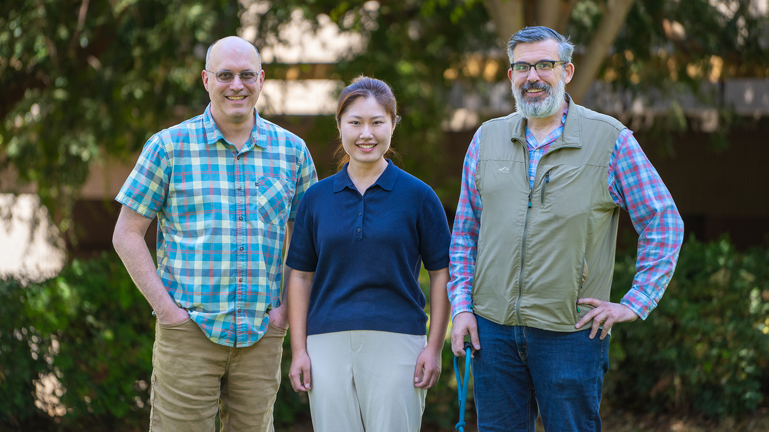Graduate student stands outdoors with two professors, all smiling in front of trees and greenery.