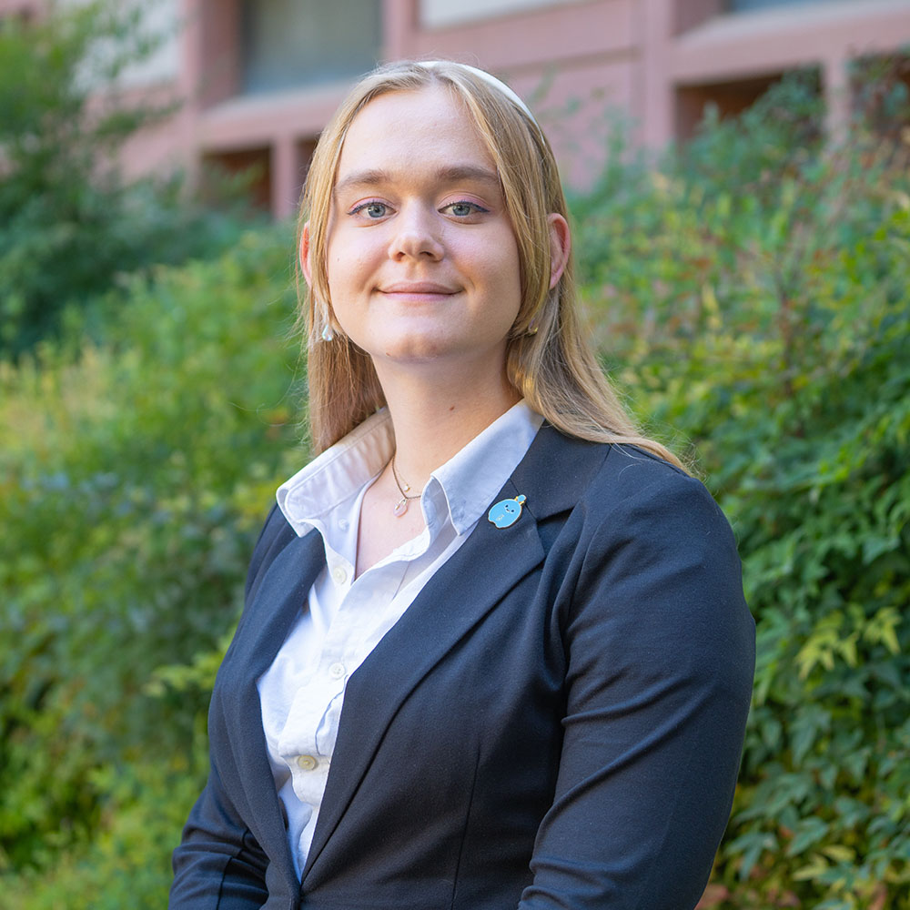 Young person with long blond hair wearing a suit jacket and white shirt, smiling outdoors in front of green bushes.