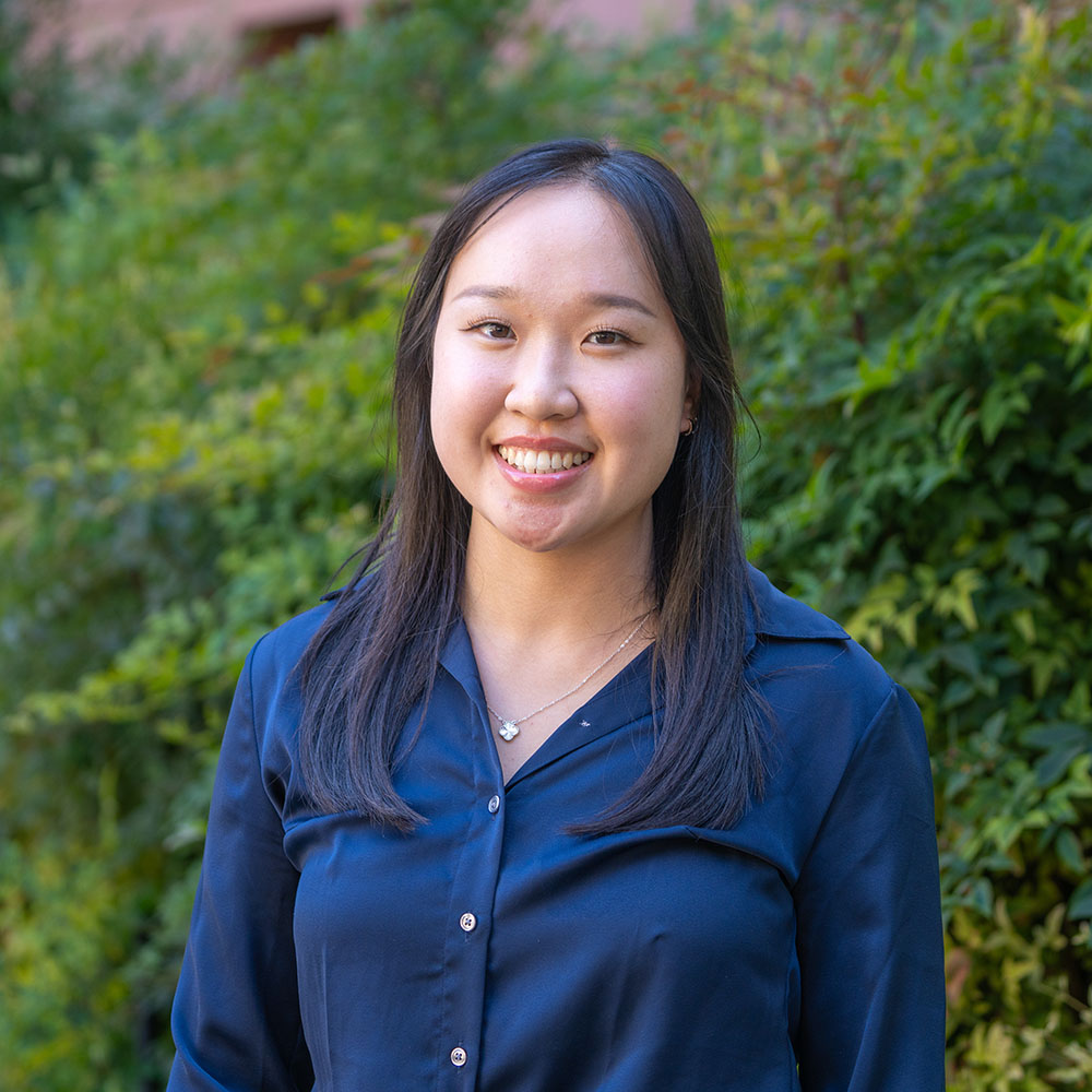ChatGPT said:  Young person with straight dark hair wearing a navy blue blouse, smiling outdoors in front of leafy green bushes.