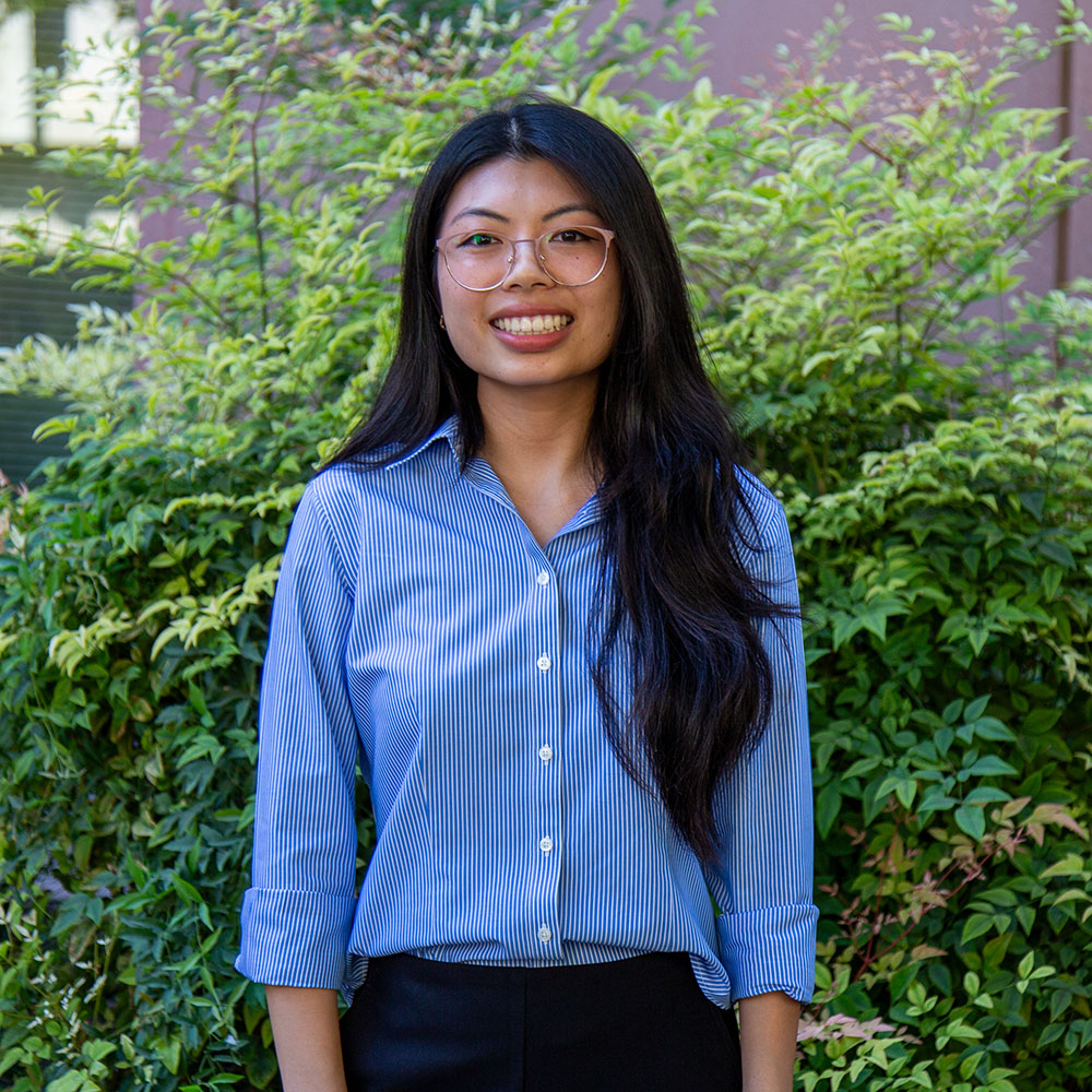 Young person with long dark hair wearing glasses and a blue striped shirt, smiling outdoors in front of green leafy bushes.