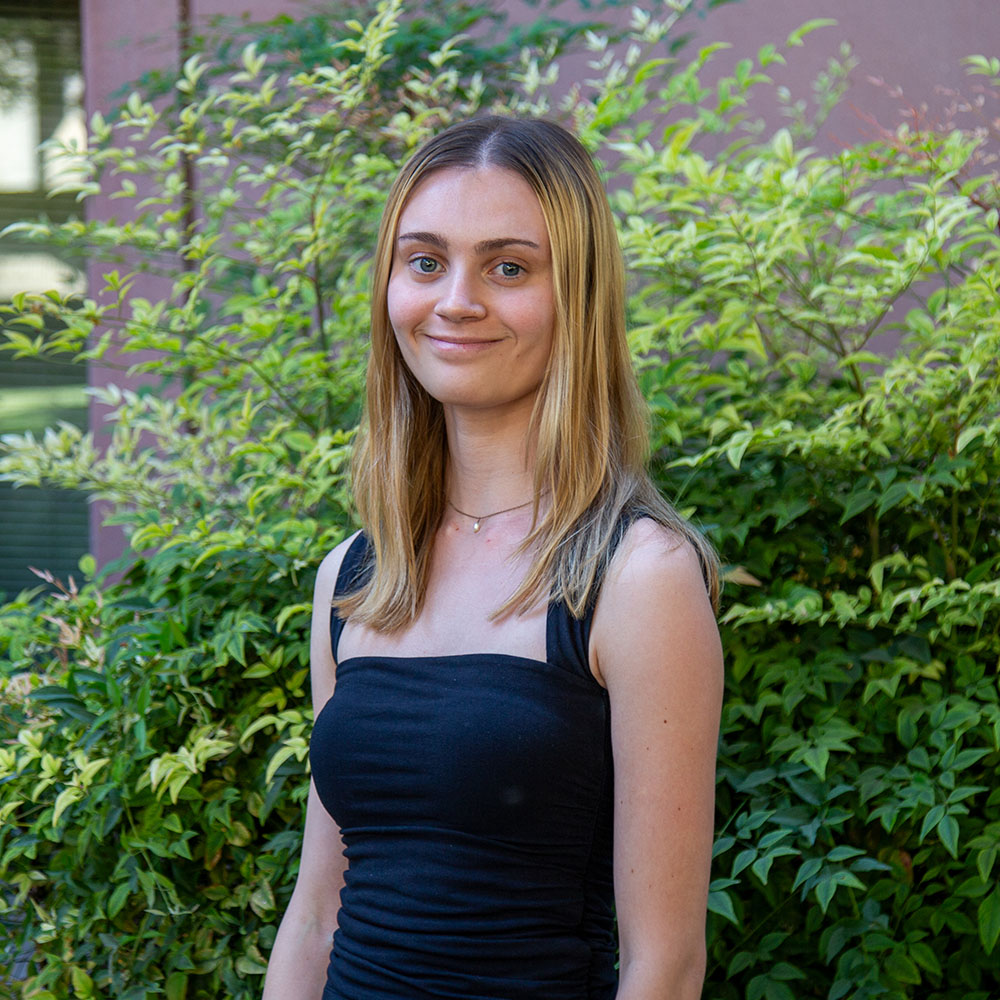 Young person with long blond hair wearing a sleeveless black dress, smiling outdoors in front of leafy green bushes.