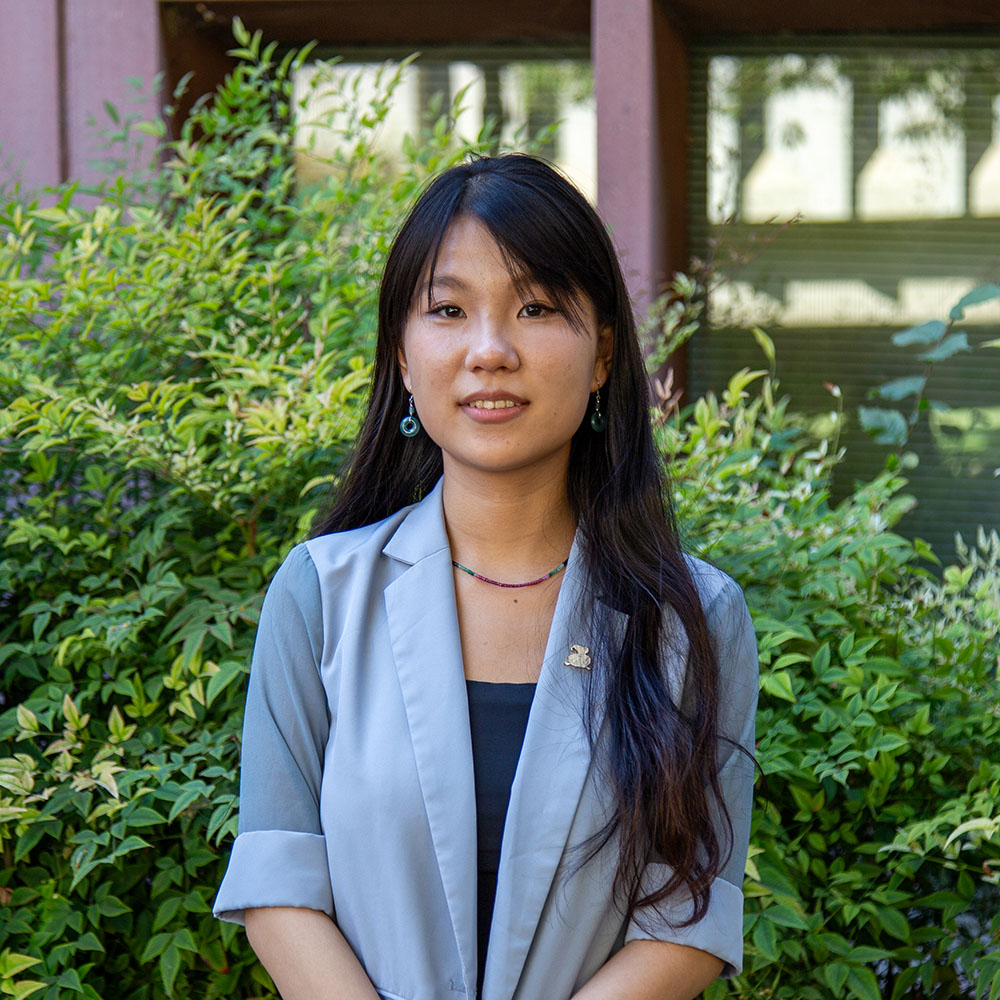 Young person with long dark hair wearing a light blazer and earrings, standing outdoors and smiling in front of green bushes.