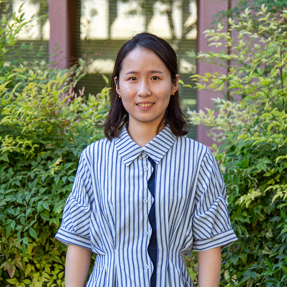 Young person with short dark hair wearing a striped button-up shirt, smiling outdoors in front of leafy green bushes.