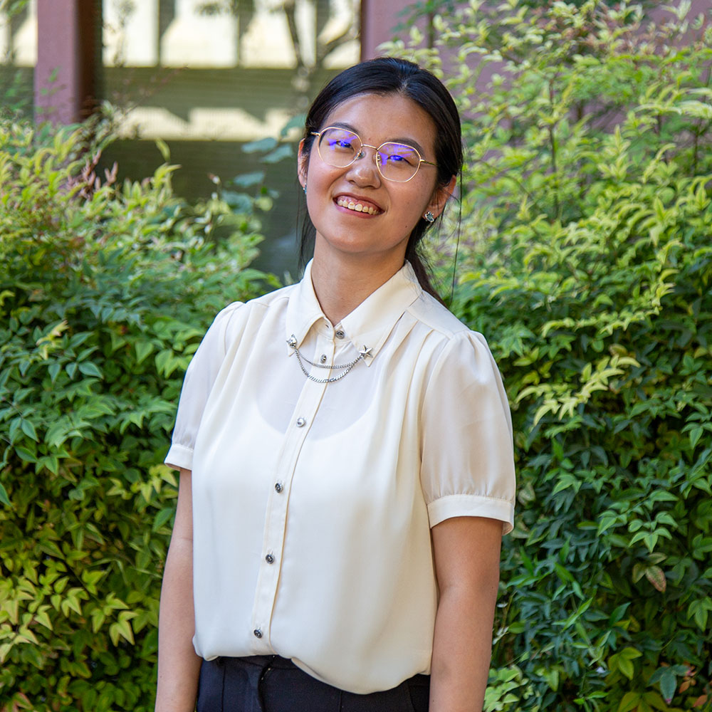 Young person with long dark hair tied back, wearing glasses and a white blouse, smiling outdoors in front of green bushes.