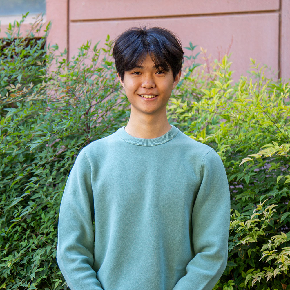 ChatGPT said:  Young person with short dark hair wearing a light green sweater, smiling outdoors in front of leafy green bushes.