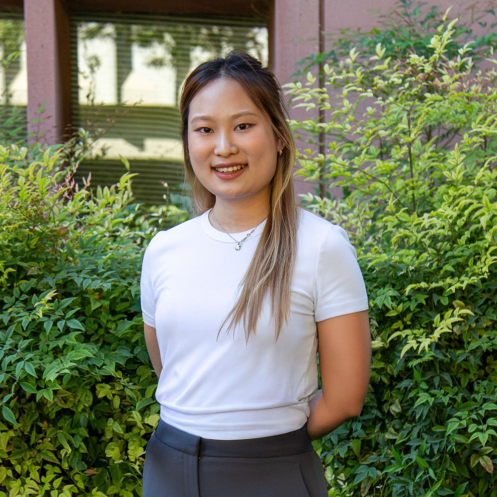 ChatGPT said:  Young person with long light brown hair wearing a white shirt and gray pants, smiling outdoors in front of green bushes.