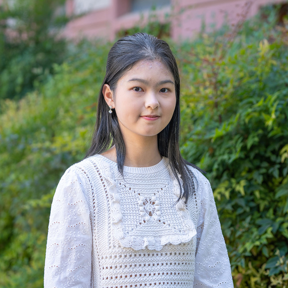 ChatGPT said:  Young person with long dark hair wearing a white crocheted blouse, standing outdoors and smiling slightly in front of green bushes.