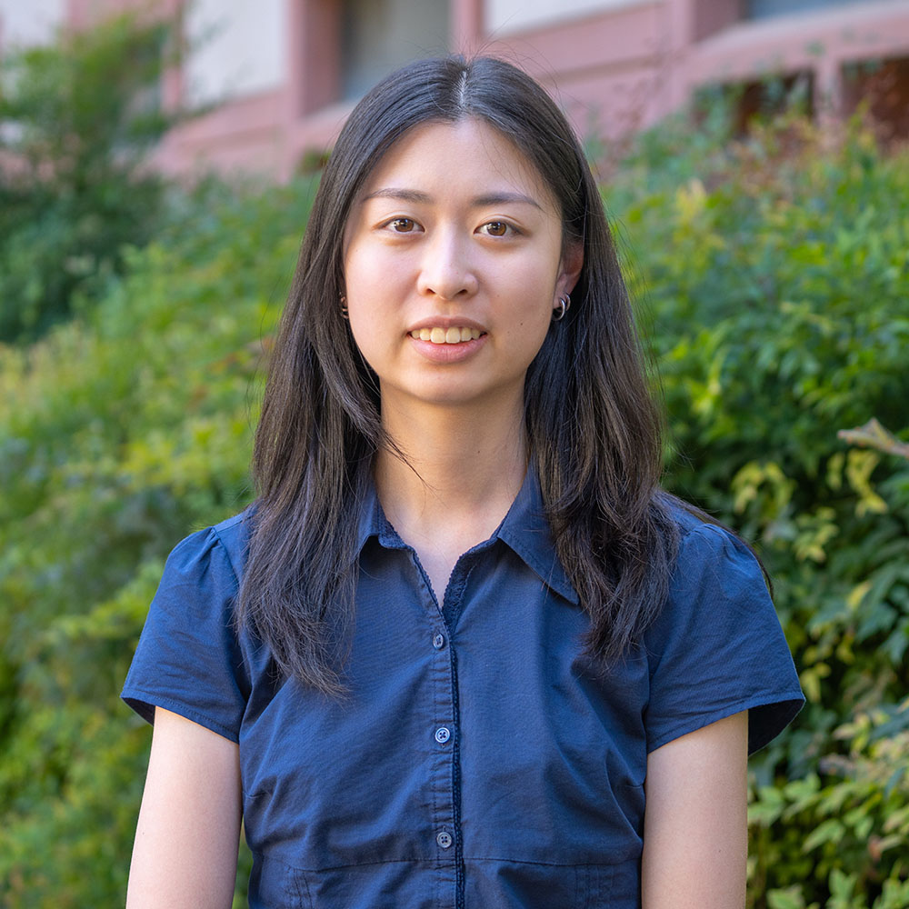 Young person with straight dark hair wearing a short-sleeved navy button-up shirt, standing outdoors and smiling in front of greenery.