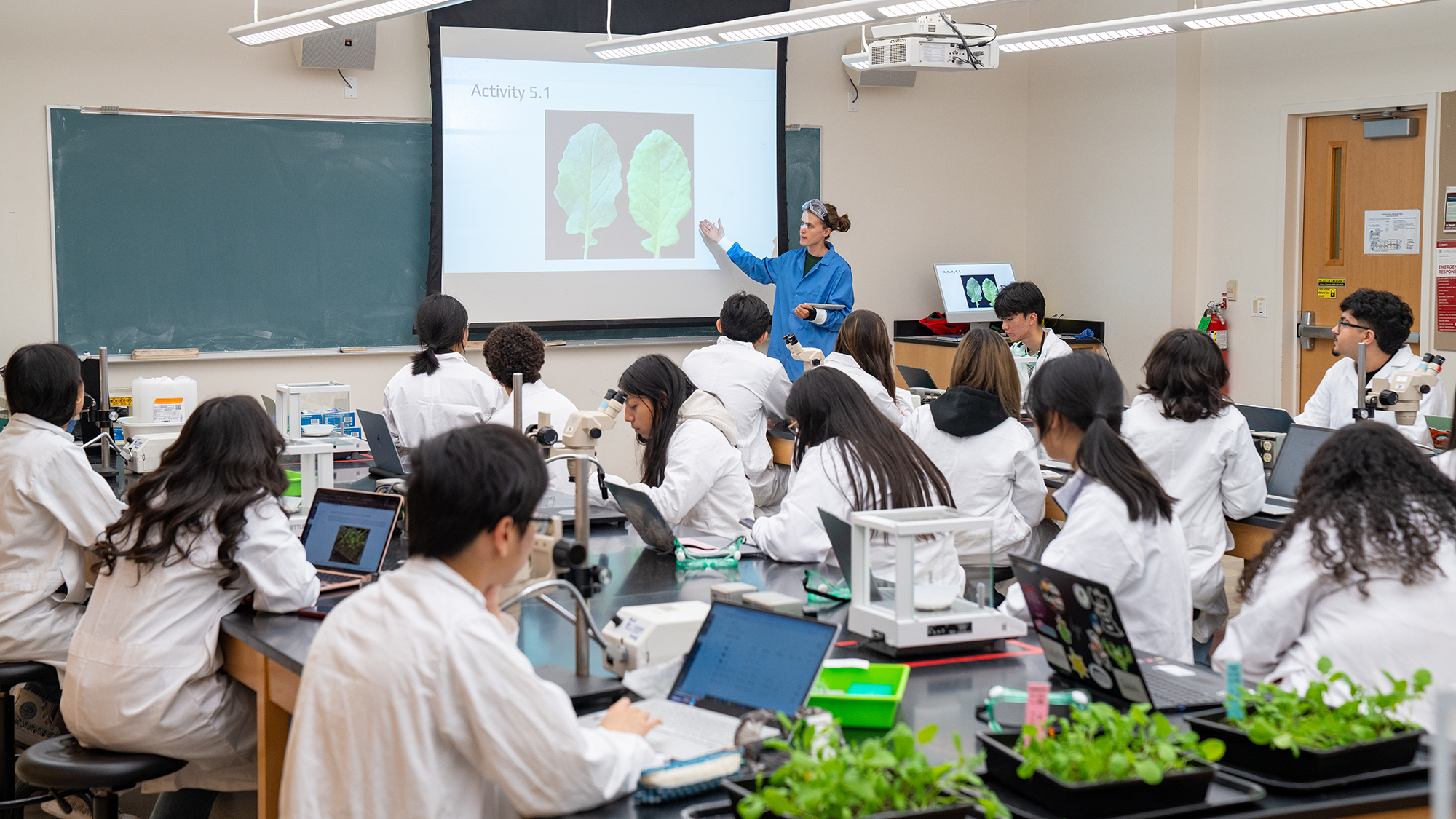 Students in lab coats listening to instructor during biology lab with leaf images projected on screen