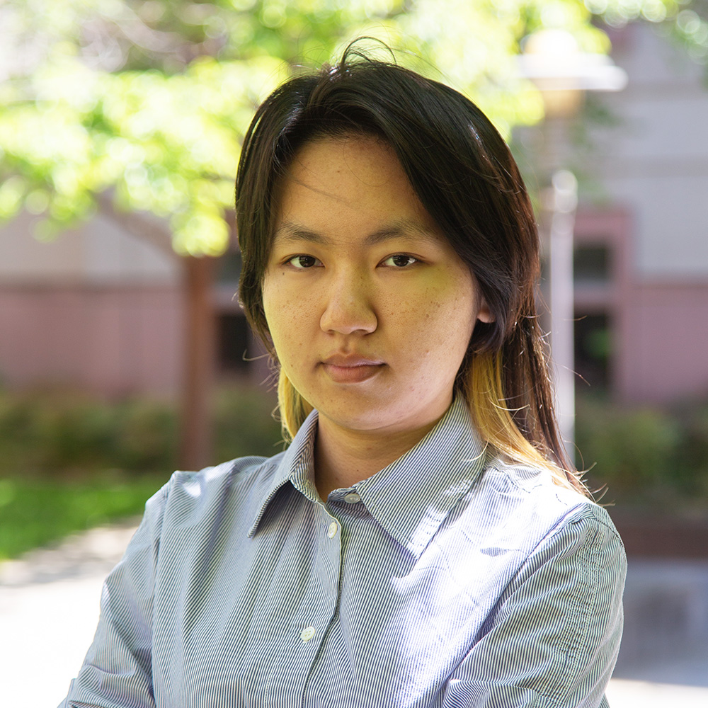 Person with dark hair in collared shirt, arms crossed, neutral expression, outdoors.
