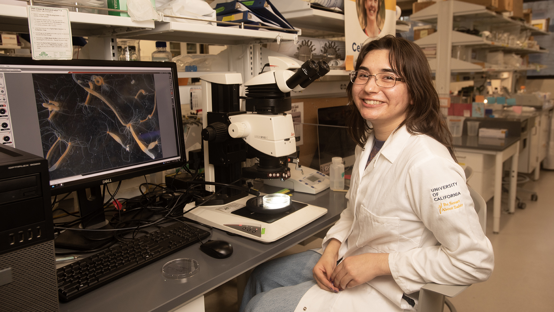 A student in a lab coat smiles while sitting at a microscope with a computer displaying images.
