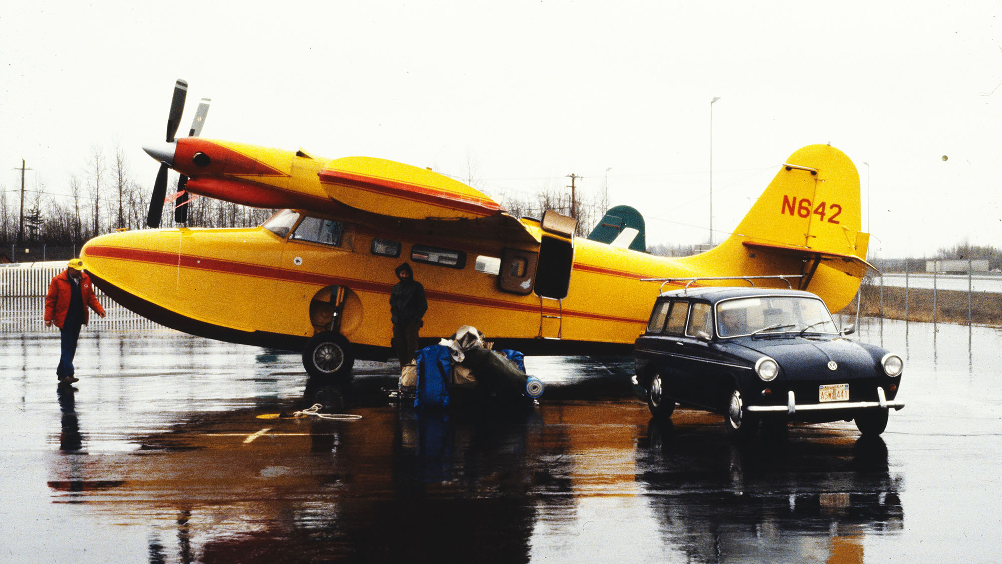A yellow seaplane on a wet runway with people and a vintage car nearby.