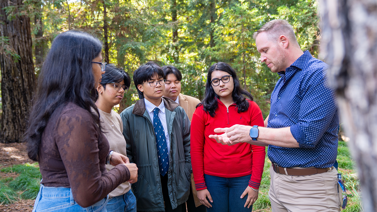 Instructor gestures to attentive diverse students standing in a forested outdoor setting.