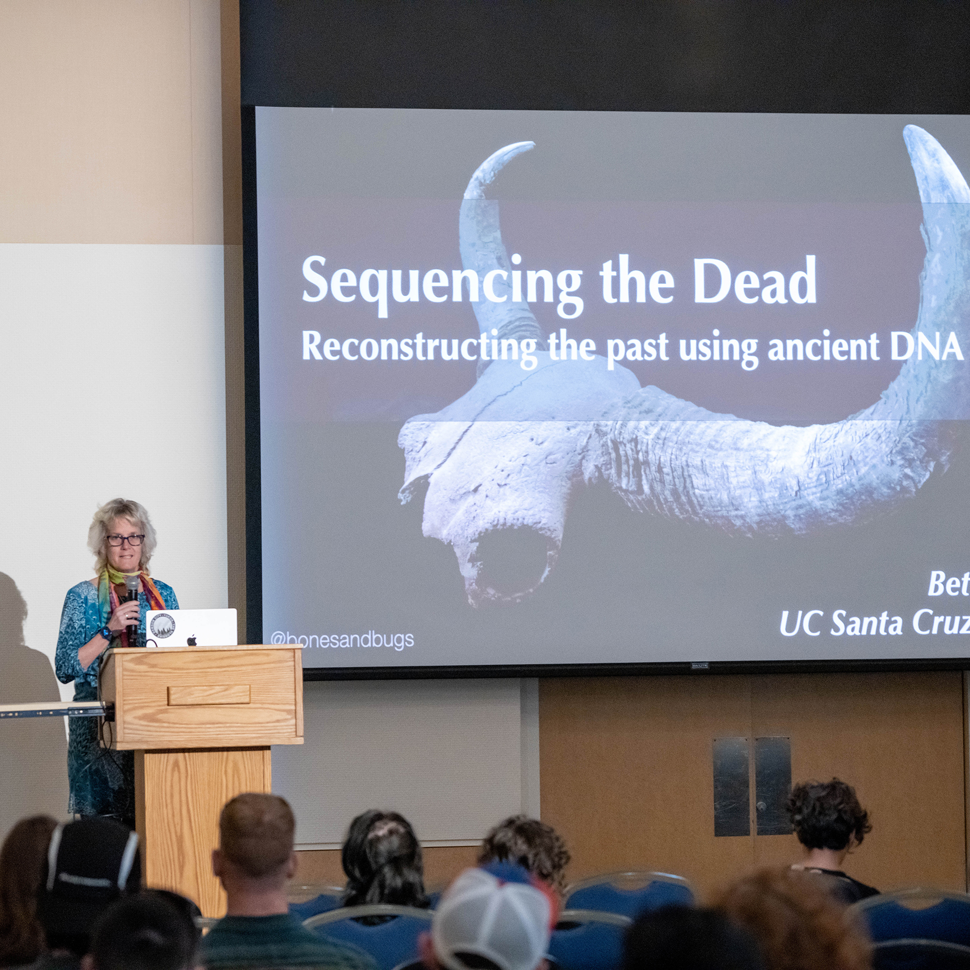 A presenter at a podium speaks about ancient DNA to an audience, with a projected slide behind.