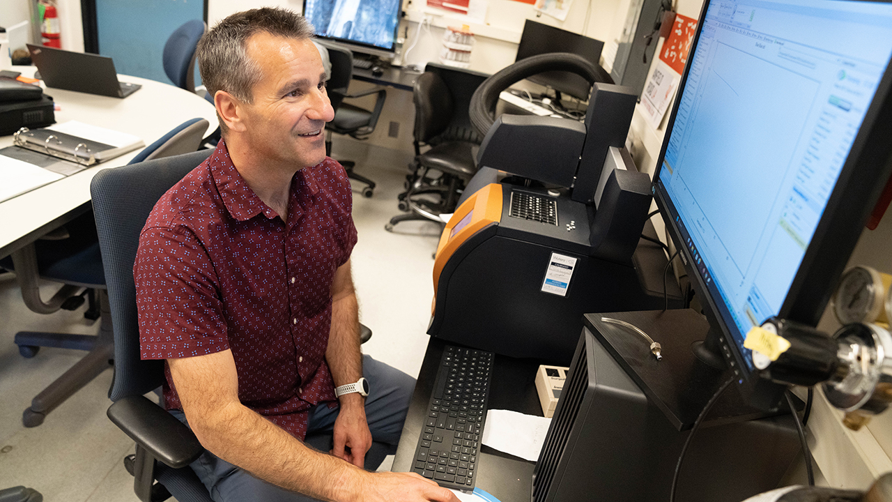 Keith Baar sits at a computer workstation in a lab, reviewing data on a monitor with scientific equipment nearby.