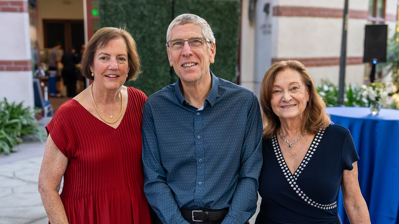 Three smiling adults outdoors; man in blue shirt between women in red and navy (photo)