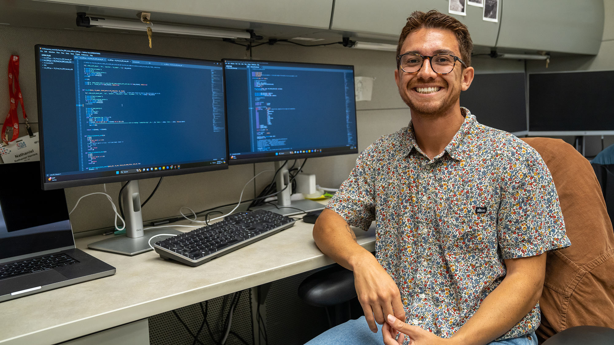 Nate Herrera, a UC Davis graduate student, smiles while seated at his workstation. Two computer monitors display coding scripts, with a keyboard and laptop on the desk. A name badge and personal touches decorate the workspace.