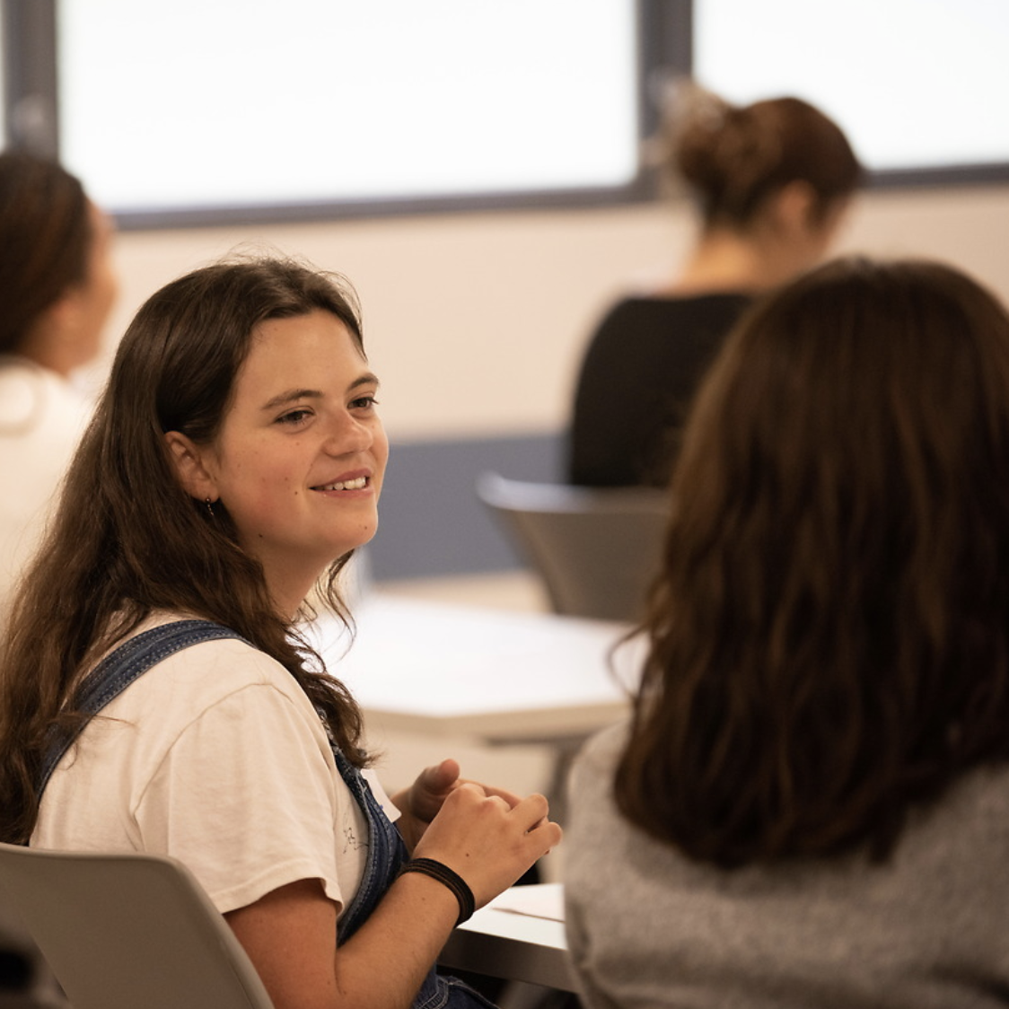Student smiling while seated in a classroom, engaged in conversation with a peer, with other students blurred in the background.
