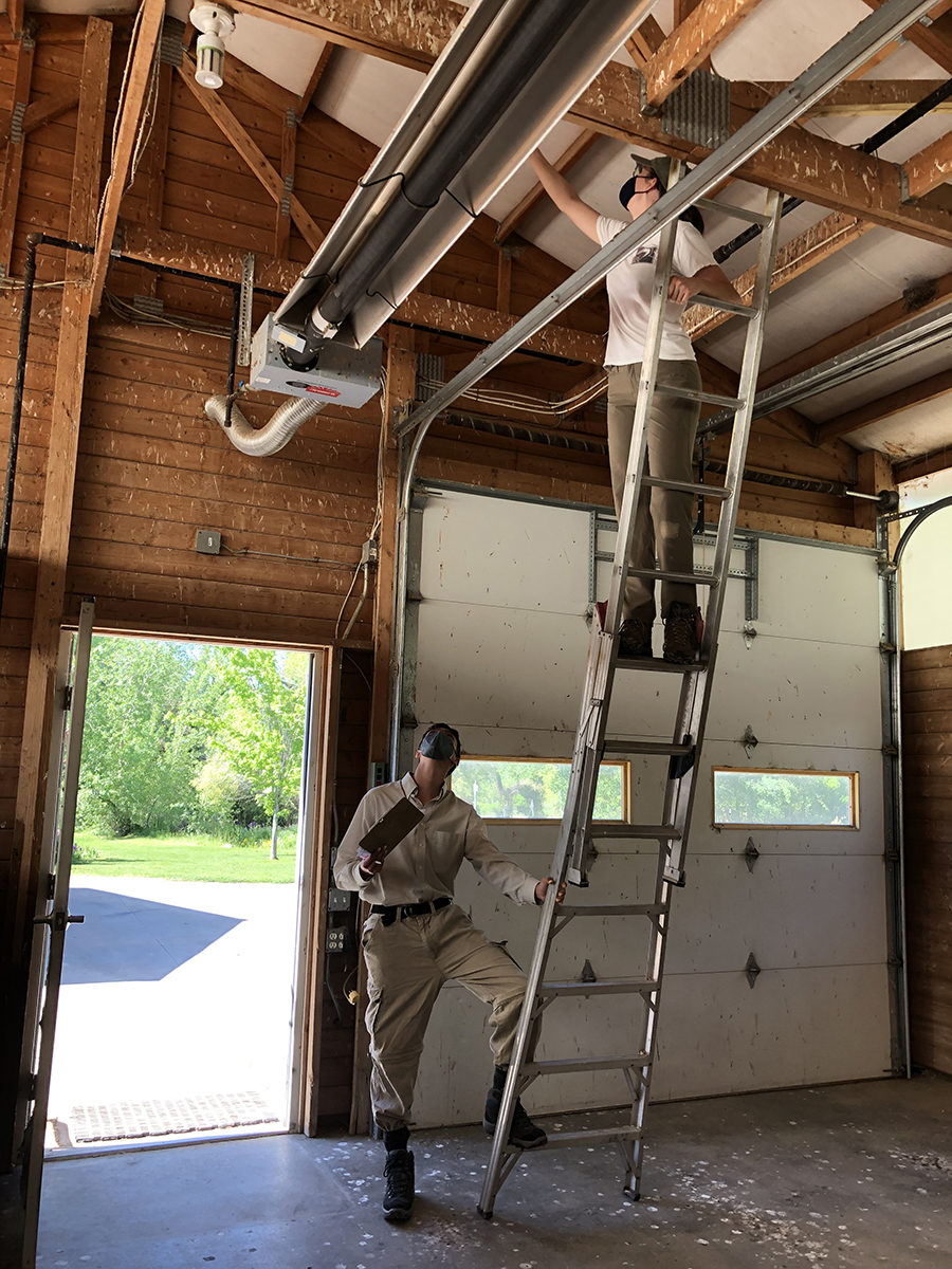 Two technicians in coveralls, one on a ladder inspecting ceiling ducts in a garage