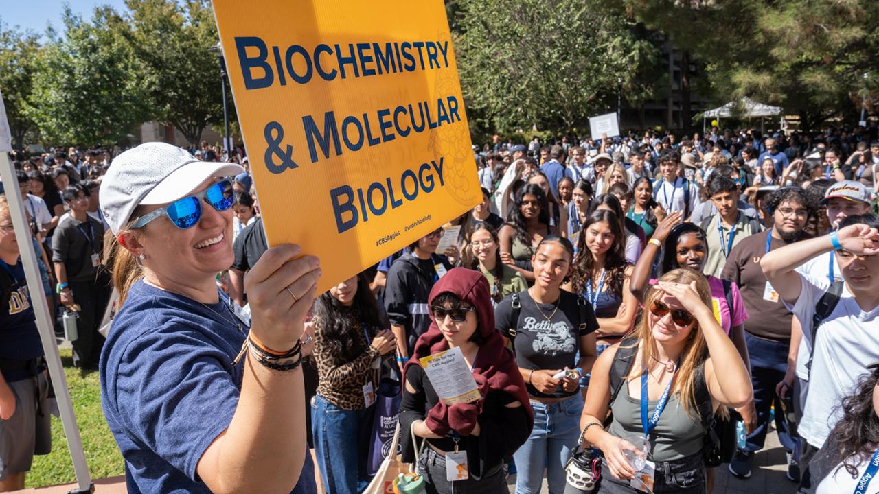 A person in sunglasses and a cap holds a yellow "Biochemistry & Molecular Biology" sign at a university orientation. Behind them, a diverse group of students watches attentively, many wearing lanyards, while trees and clear skies frame the outdoor event.