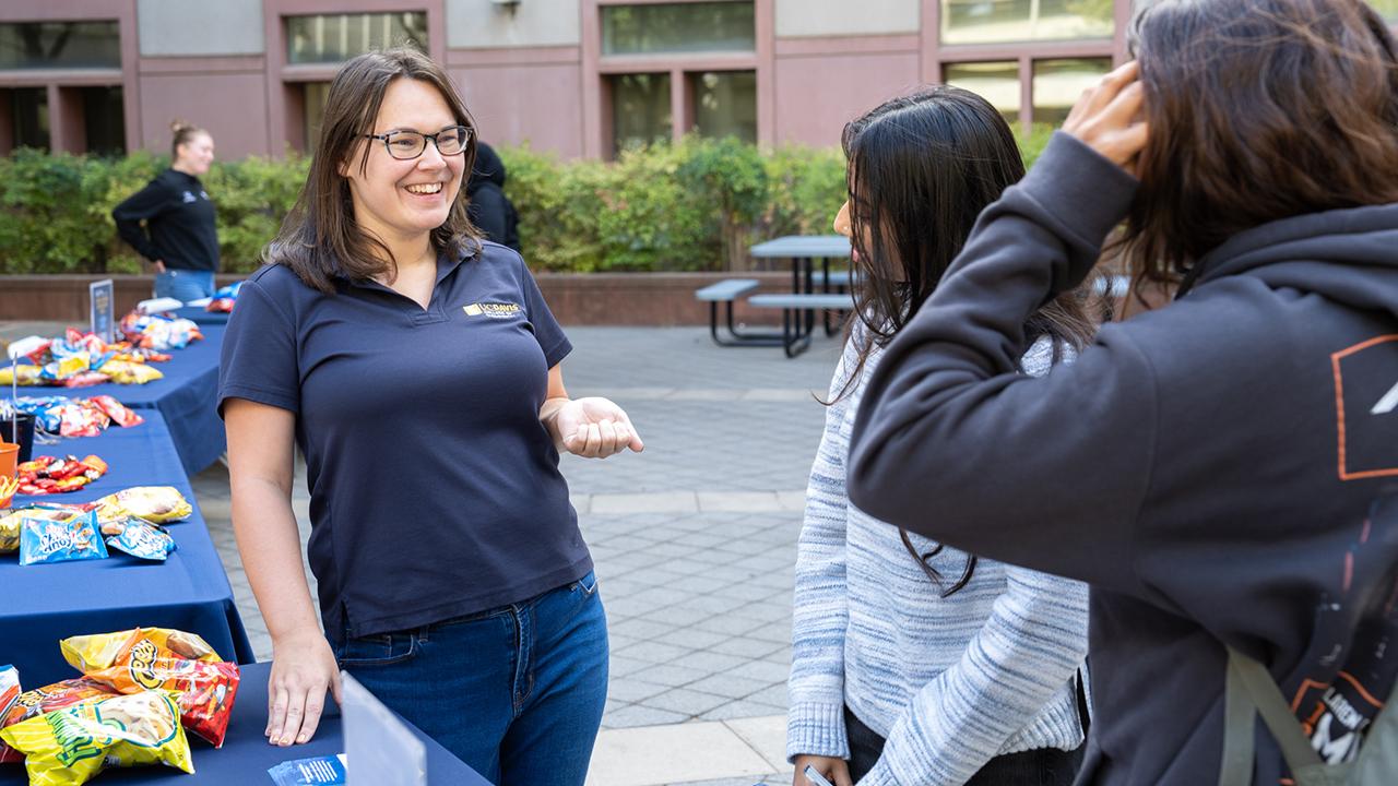 A friendly UC Davis staff member wearing a UC Davis polo shirt engages in conversation with two students at an outdoor information fair. Tables covered with snacks are visible in the background.