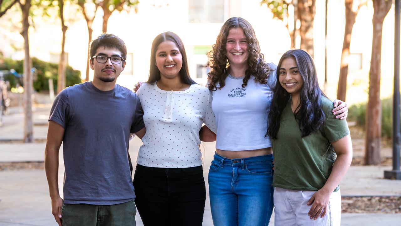 Four young adults stand side by side outdoors with their arms around each other, smiling warmly at the camera. The group consists of two men and two women, all casually dressed and appearing relaxed and friendly. Sunlight filters through trees in the background, casting a gentle glow on the group, who are standing on a paved path with greenery and a building partially visible behind them.
