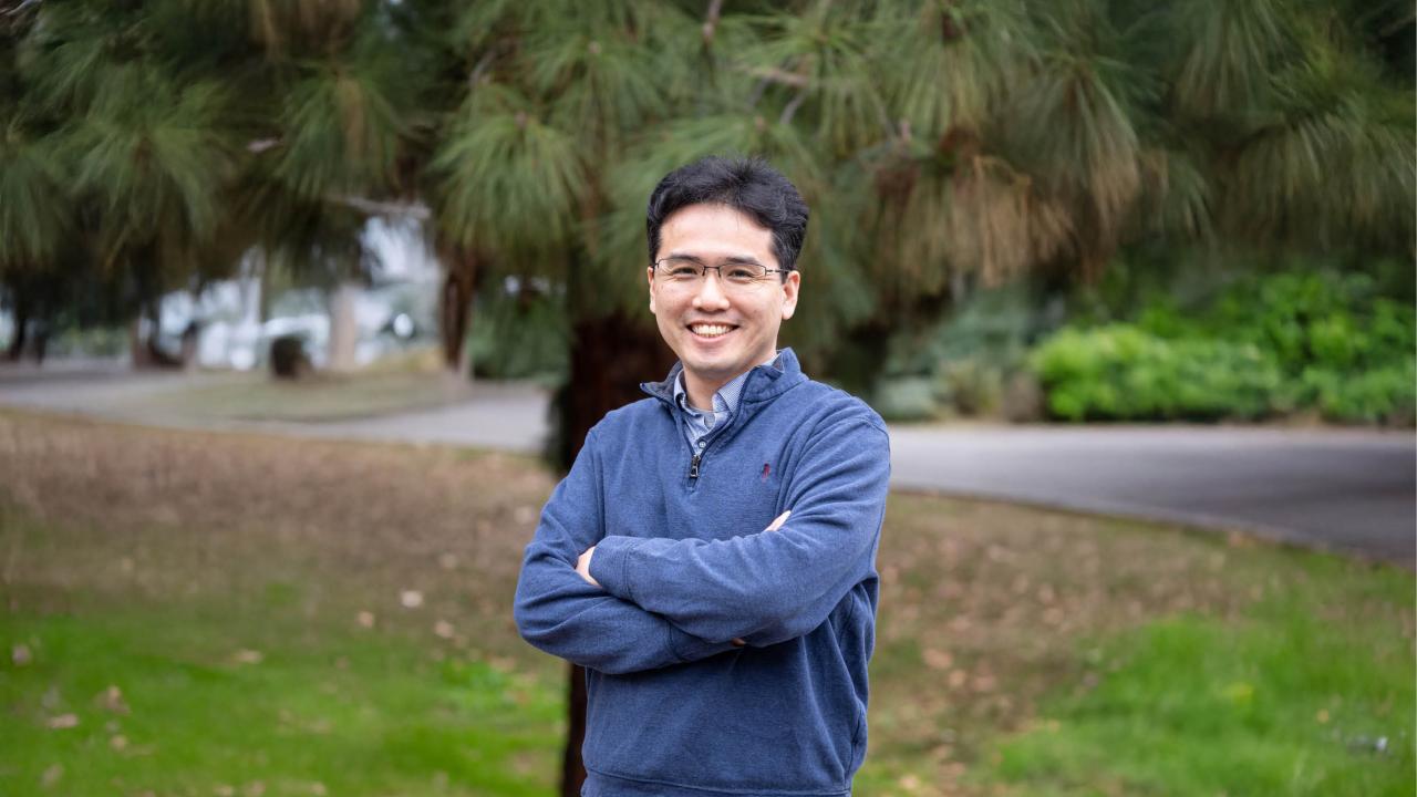 A man wearing glasses and a blue pullover stands outdoors with his arms crossed, smiling at the camera. He is positioned in front of a backdrop of pine trees and a curved path, with green grass and scattered leaves in the foreground.