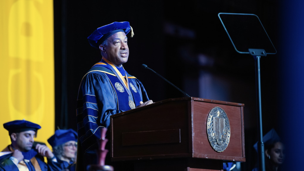 A university official in blue and gold academic regalia delivers a speech at a graduation ceremony, standing behind a podium with a university seal, while graduates listen in the background.