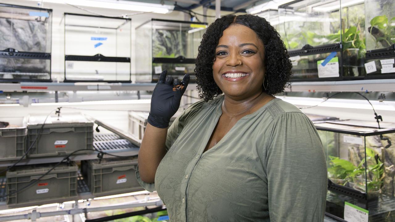 A smiling woman wearing gloves holds a small crab in a laboratory filled with terrariums and plant enclosures.