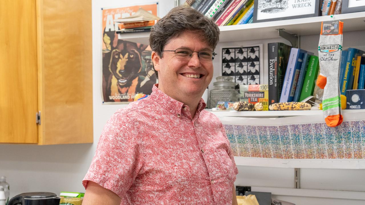 A smiling man wearing glasses and a red patterned shirt stands in a colorful office or lab space. Behind him are shelves filled with books, framed artwork, and quirky items.
