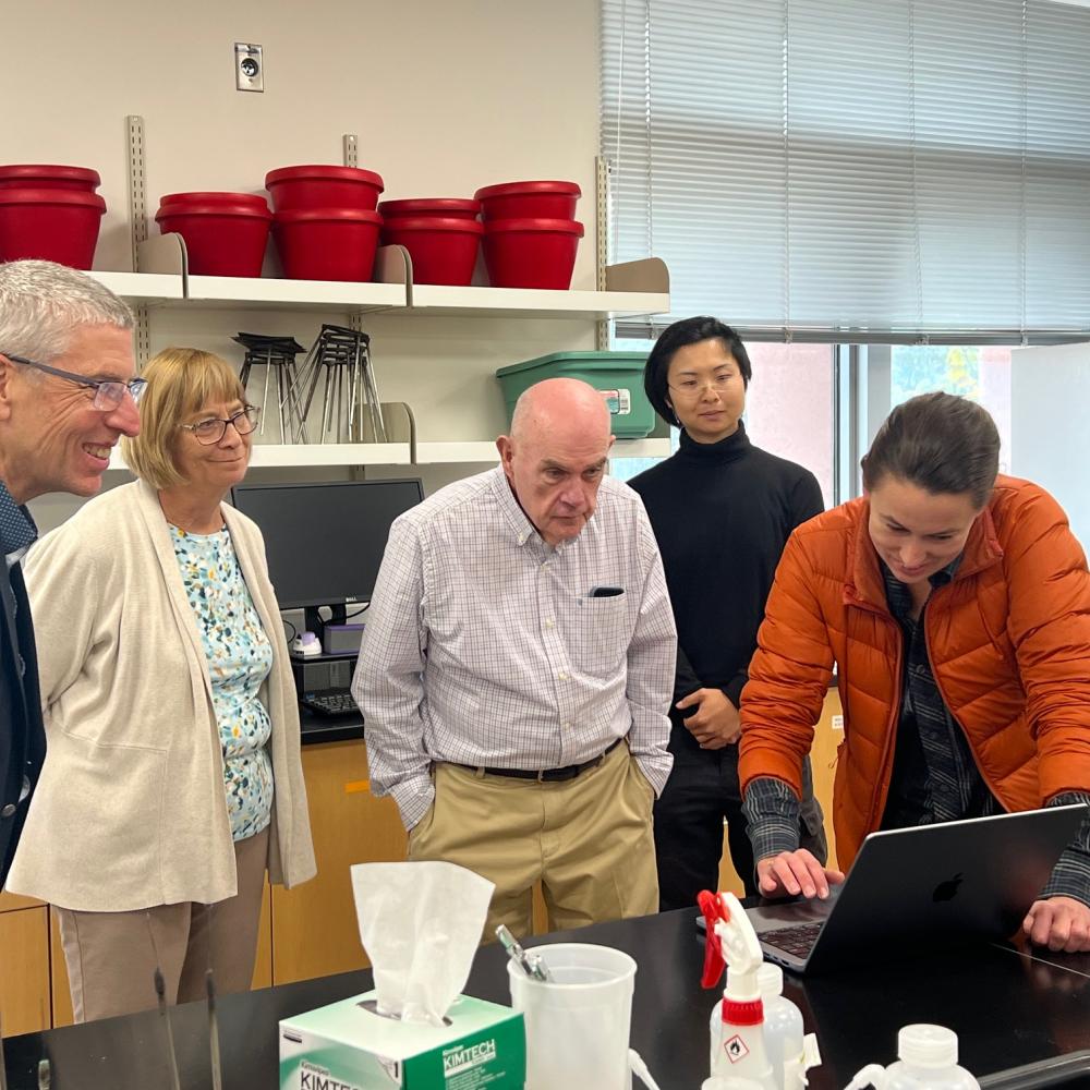 A small group of visitors gathers around a UC Davis lab bench as a researcher shows data on a laptop.