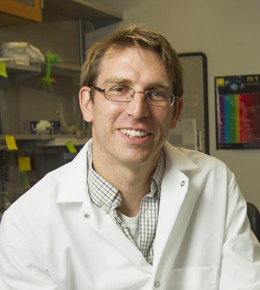 A smiling man wearing glasses, a white lab coat, and a plaid shirt, seated in a laboratory setting with scientific equipment and colorful sticky notes in the background.