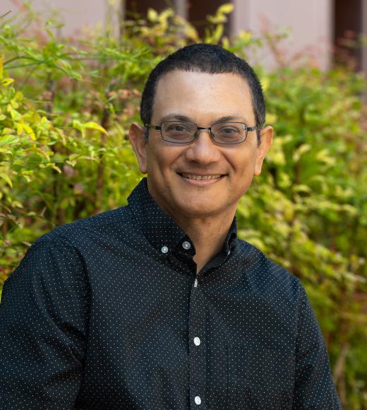 Portrait of a smiling person with short hair and glasses wearing a black button-up shirt with white polka dots, standing in front of green foliage.