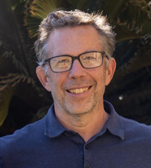 A headshot of a man with short grey-black hair, glasses and a blue shirt standing in front of a palm tree.