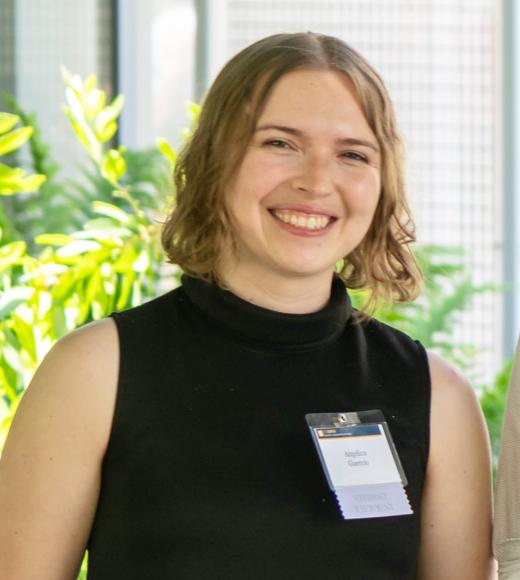 A smiling person with short, wavy light brown hair wearing a sleeveless black turtleneck top. They are standing indoors with green plants in the background and wearing a name badge that reads “Angelica Guerrero” with a ribbon labeled “Student Recipient.”
