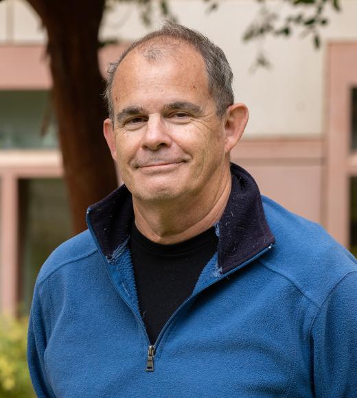 Portrait of a man with short gray hair, wearing a blue fleece jacket over a black shirt, standing outdoors with a building and trees in the background. He has a slight smile and is looking directly at the camera.