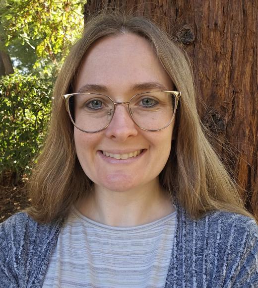 Smiling person with glasses and long hair stands in front of a large tree, wearing a striped shirt and gray cardigan.