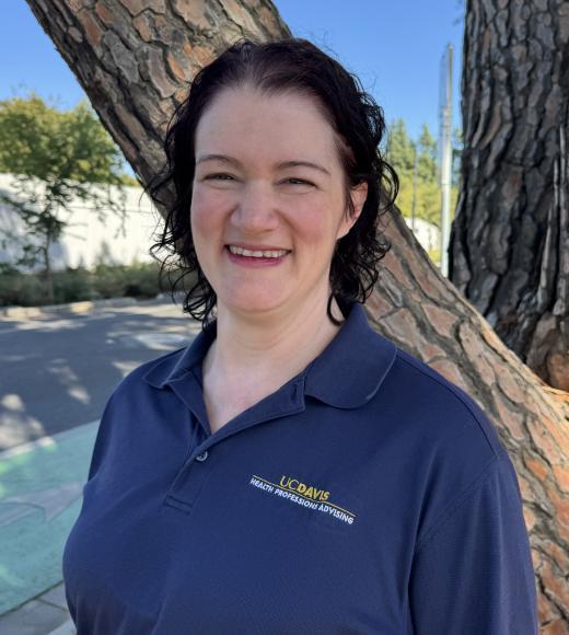 Smiling woman with dark hair in navy polo leaning against tree outdoors