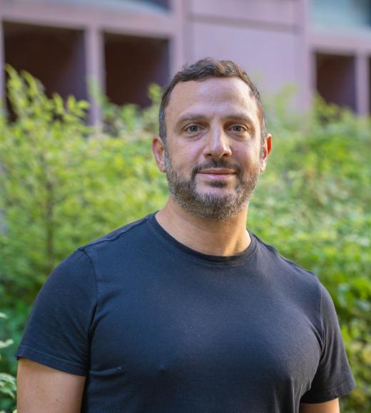Person standing outdoors, wearing a black t-shirt, short dark hair and beard, greenery and building behind.