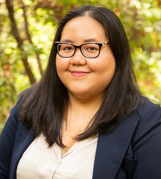 Smiling woman with glasses, dark hair, navy blazer, leafy outdoor background.