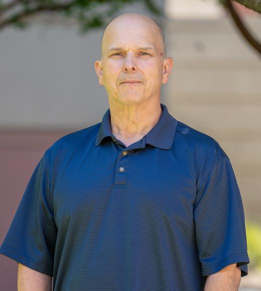 Outdoor portrait photograph of a bald man in a navy polo, neutral expression