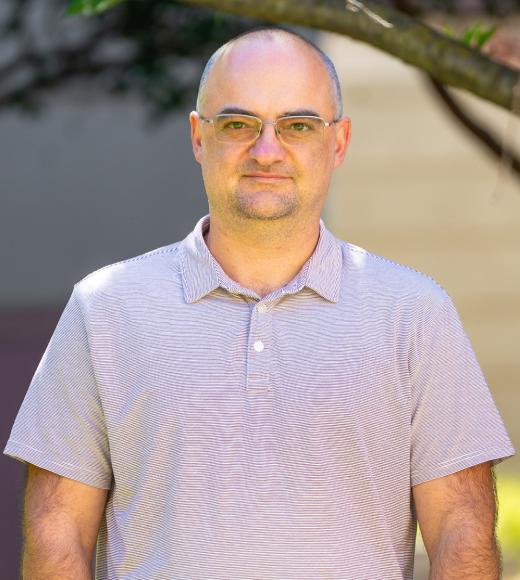 Headshot of bald man in glasses wearing lavender polo, neutral expression, blurred greenery
