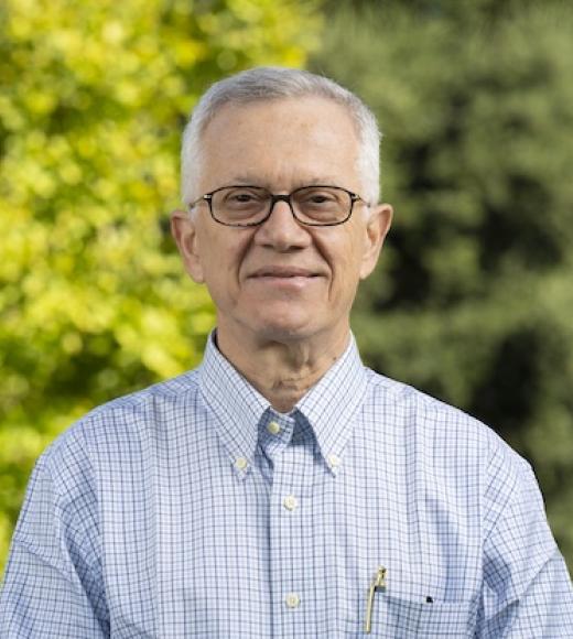 Smiling person with short gray hair and glasses wears a light blue plaid shirt, standing outdoors with trees in the background.