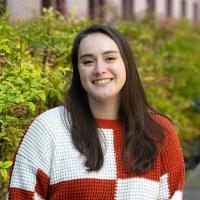 Portrait of a smiling person with straight dark brown hair, wearing a red and white checkered sweater, standing in front of green foliage.