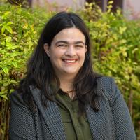 Portrait of a smiling person with long dark hair and a nose ring, wearing a gray blazer over a dark green shirt, in front of green shrubbery.